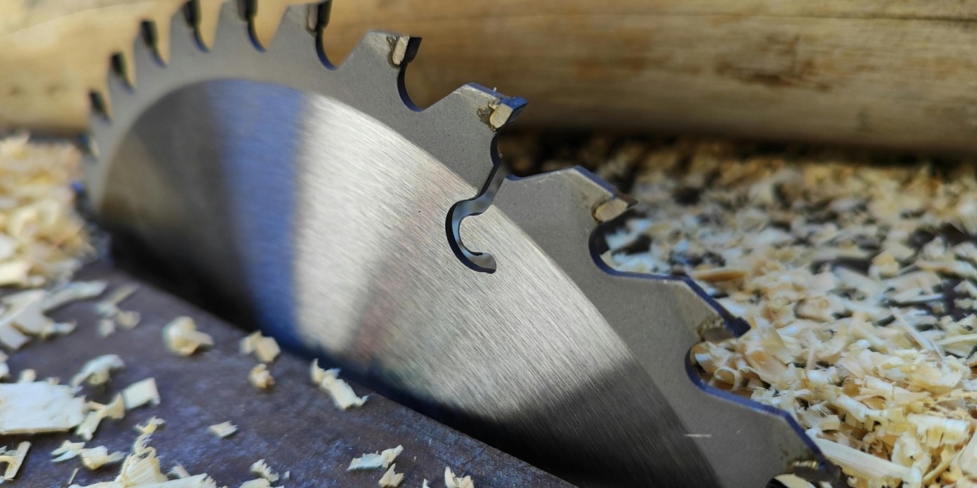Hoja de sierra circular parcialmente incrustada en una mesa de trabajo, rodeada de virutas de madera, con troncos en el fondo.